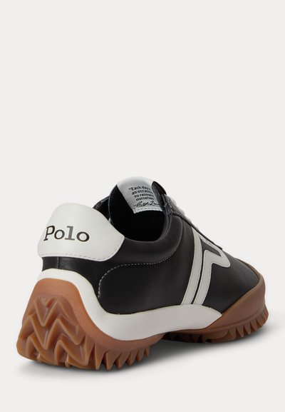Μαύρο και άσπρο αθλητικό παπούτσι Polo με καφέ ζιγκ ζαγκ σόλα, λευκή πλαϊνή ρίγα και λογότυπο Polo στην ταμπελίτσα της φτέρνας, λοξή όψη που δείχνει την εσωτερική ετικέτα.