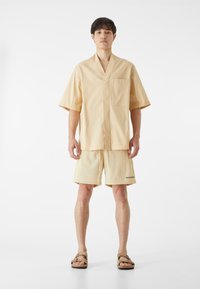 Pale žltý bavlnený loungewear set pozostávajúci z trička s krátkym rukávom bez goliera a zodpovedajúcich krátkych nohavíc s elastickým pásom a detailom loga.