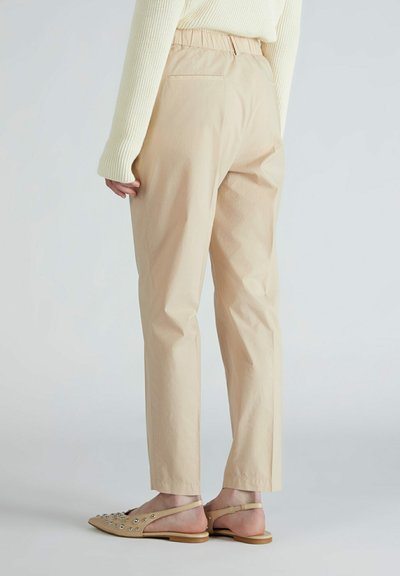 Pantalons beiges taille haute à coupe décontractée, dotés d'une poche arrière et associés à des sandales à semelles plates et ouvertes ornées de clous en argent.