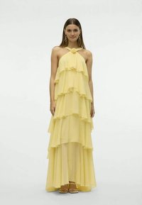 Donna che indossa un maxi abito giallo pallido senza maniche, a più livelli, con un dettaglio floreale sul colletto e sandali beige su uno sfondo neutro.