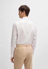 Chemise blanche à manches longues et col pointu, portée avec un pantalon en laine beige clair présentant une texture subtile et des poches arrière.
