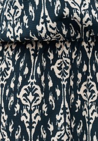 Tissu bleu sarcelle foncé et blanc cassé avec un motif floral et de vigne symétrique et complexe, montrant des plis et la texture du matériau tissé.