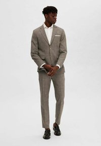 Costume à carreaux gris avec une coupe ajustée, veste à un bouton, et pantalon fuselé assorti, accompagné d'une chemise blanche et de chaussures noires.