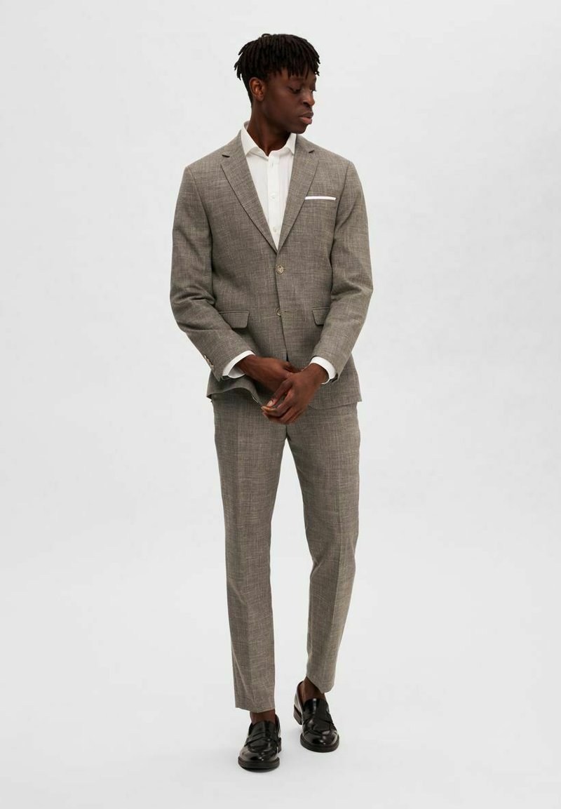 Costume à carreaux gris avec une coupe ajustée, veste à un bouton, et pantalon fuselé assorti, accompagné d'une chemise blanche et de chaussures noires.