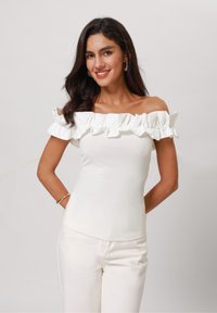 Off-shoulder valkoinen paita, jossa on röyhelökaulus, kuvioitu pinta ja tiivis muotoilu. Yhdistetty vaaleanvärisiin housuihin.