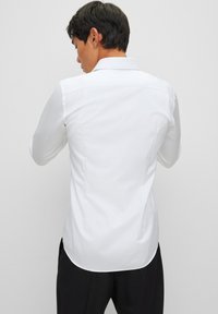 Camisa blanca de manga larga con cuello puntiagudo, que presenta una textura suave, un ajuste entallado y un dobladillo curvado. La vista trasera muestra un sutil detalle de costuras.