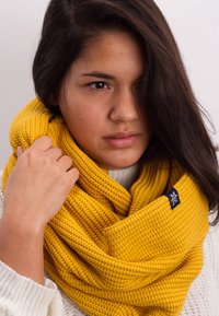 Yellow knitted infinity scarf with a textured pattern, wrapped around the neck, featuring a small black logo tag on the side.