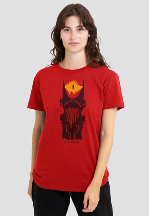 Woman wearing red t-shirt with black and yellow Eye of Sauron helmet graphic and "Lord of the Rings" text, standing against gray background.