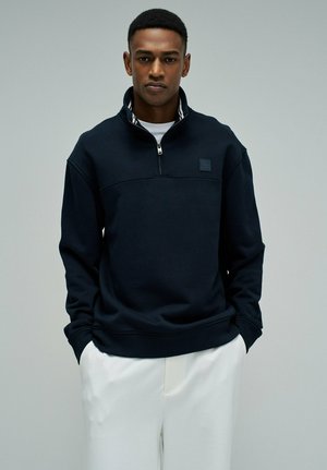 Hombre con un suéter negro de media cremallera y manos en los bolsillos de pantalones blancos, frente a un fondo gris liso.