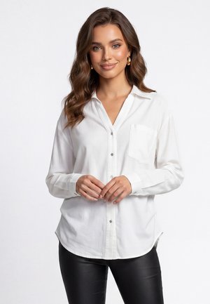 Donna con capelli castani mossi, che indossa orecchini a cerchio dorati, una camicia bianca abbottonata e pantaloni neri, in piedi con le mani delicatamente intrecciate.