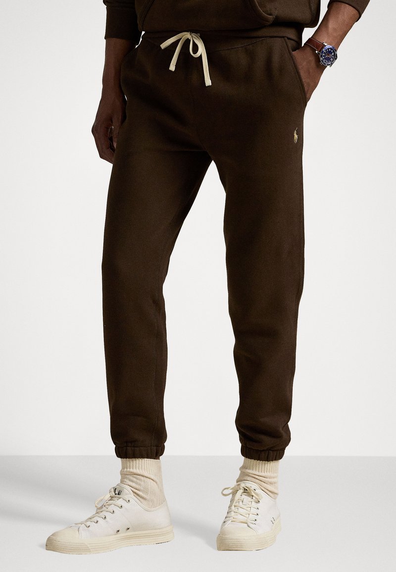 Brune joggebukser med snor i livet, ribbestrikkede mansjetter og sidelommer; har et lite logo på venstre lår og er sammen med hvite joggesko.