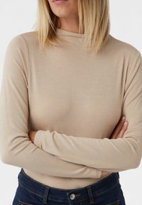 Beige Langarmshirt mit hohem Kragen und gerippter Textur, getragen mit dunklen Jeans, die sichtbare Nähte und einen sichtbaren Knopfdetails aufweisen.