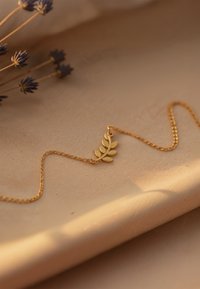 Sart guld kæde med en bladformet vedhæng hvilende på blød beige stof, tørrede lilla blomster i baggrunden.