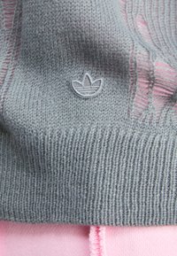 Grå stickad tyg med ribbat mönster, med en liten grå Adidas-logotyp. Nere, ljusrosa tyg synligt.