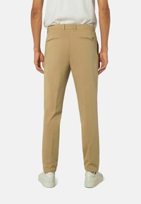 Mann in beigen, schmal geschnittenen Hosen und weißen Sneakers, der mit dem Rücken zur Kamera vor einem schlichten Hintergrund steht.