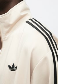Krem sportska jakna s visokim ovratnikom, crnim detaljima od tri pruge na rukavima i malim crnim Adidas logotipom na prsima.