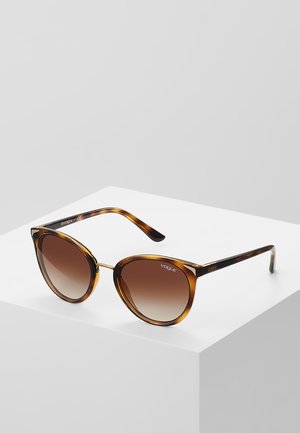 Kattens oog zonnebril met een schildpadmotief, verloopbruin lenzen en een sleek zwart-gouden logo op de pootjes.