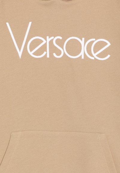 Beige bomullshuvtröja med en framficka och en framträdande vit "Versace"-logotyp över bröstet. Slät textur, normal passform.