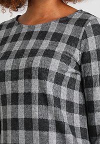 Femme portant une chemise à manches longues à carreaux gris et noirs avec un col rond, présentant un motif de tissu texturé.