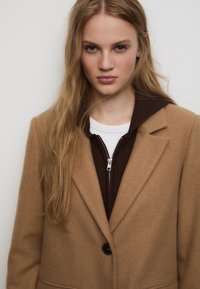 Cappotto in lana marrone con colletto a punta e un bottone nero, indossato sopra un maglione marrone scuro con zip e una camicia bianca.