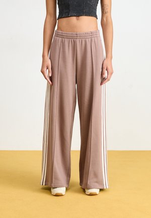 Persona que lleva pantalones marrones de corte holgado con rayas blancas en los lados, zapatillas blancas y una camiseta negra corta, de pie sobre un suelo amarillo.