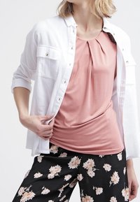 Biała koszula z guzikami noszona na różowej bluzce z plisami, w zestawieniu z czarnymi spodniami w kwiaty, na których znajdują się różowe i białe kwiaty.