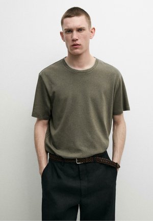 Młody mężczyzna z krótkimi włosami, ubrany w oliwkowo zieloną koszulkę, czarne spodnie i pleciony brązowy pasek, stojący na jednolitym jasnym tle.