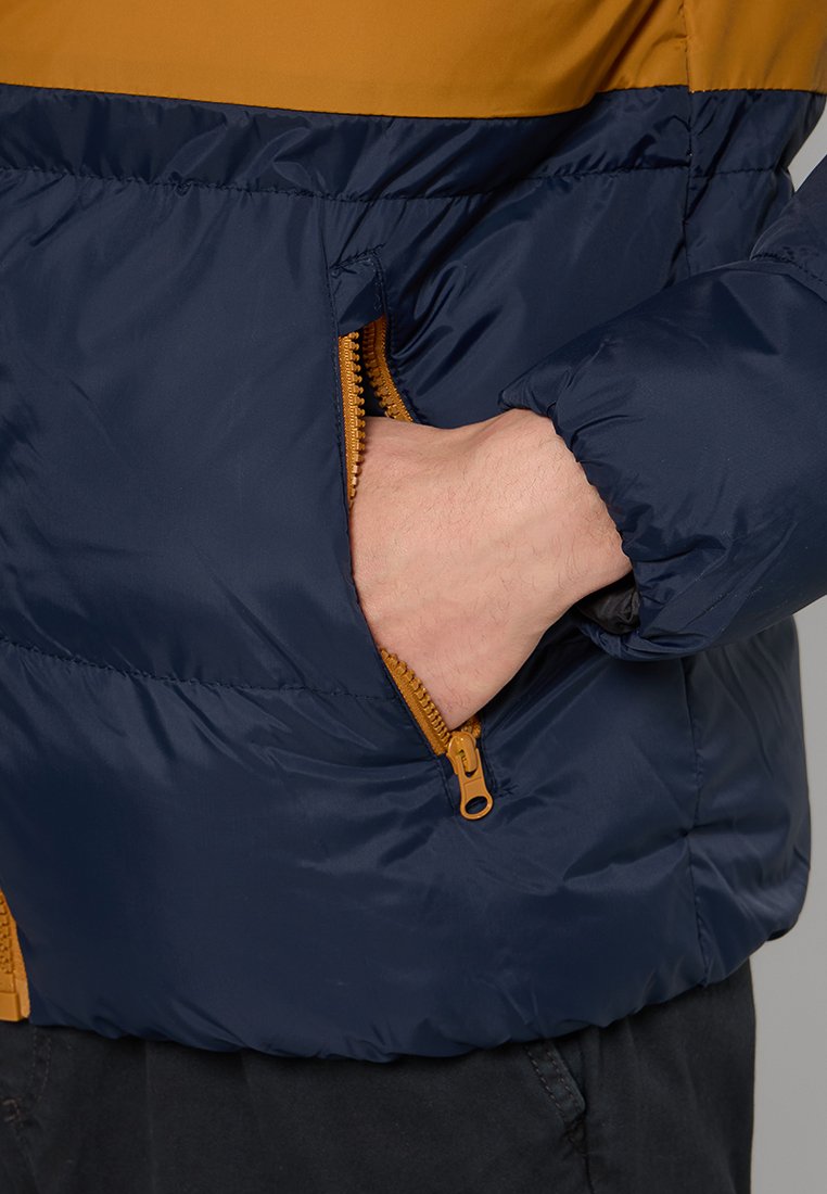 Navy blauwe pufferjack met een mosterdgele bovenkant, voorzien van een zichtbare ritszak en elastische manchetten. Glad, glanzend textuur.