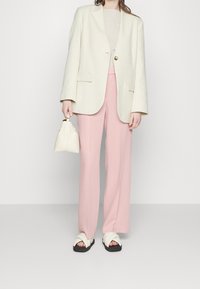 Krämfärgad blazer med guldknapp, ljusbeige topp, mjuka rosa vida byxor, vit väska och vita snurrsandaler med svarta sulor.