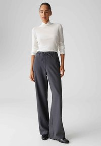 Grijze broek met trekkoord en losse pasvorm, gemaakt van gestructureerde stof. Gecombineerd met een wit shirt met lange mouwen en een hoge kraag.