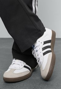 Białe buty sportowe z szarymi zamszowymi akcentami, trzema czarnymi paskami po bokach i brązową gumową podeszwą, noszone z czarnymi spodniami.