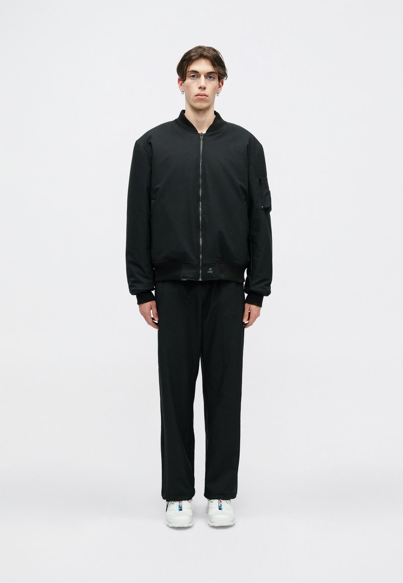 Melnā bomber jaka ar rāvējslēdzēju priekšpusē un ribotām piedurknēm, apvienota ar vaļīgiem melniem biksēm un baltām sporta apaviem. Minimalistisks dizains.