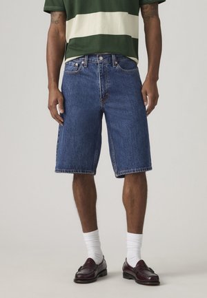 Mand iført blå denimshorts, grøn- og hvidstribet skjorte, hvide sokker og mørkebrune loafers, stående foran en neutral baggrund.