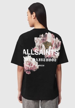 Kvinde med langt mørkt hår iført en sort AllSaints t-shirt med store lyserøde og hvide blomstergrafikker på ryggen.