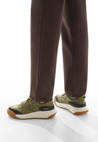 Sneakers verde oliva con tessuto testurizzato, punte rinforzate, accenti neri e suola in gomma crema. Abbinati a pantaloni marrone scuro.