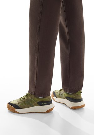 Sneakers verde oliva con tessuto testurizzato, punte rinforzate, accenti neri e suola in gomma crema. Abbinati a pantaloni marrone scuro.