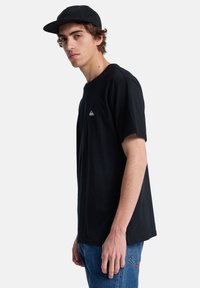 Camiseta de algodón negra con mangas cortas, cuello redondo y un pequeño logo blanco en el lado izquierdo del pecho. Llevada con vaqueros de denim azules.