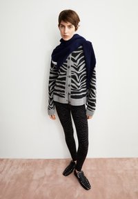 Cardigan à rayures zèbres gris et noir, leggings noirs à motif zèbre, écharpe marine drapée sur les épaules et chaussures à enfiler noires à clous.