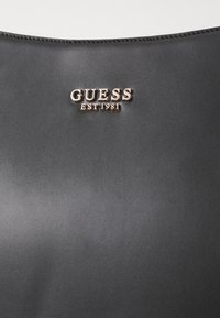 Melnas sintētiskas ādas rokas somas, kuras priekšpusē ir izcils zelta "GUESS EST 1981" logotips, gluda tekstūra bez papildus rakstiem.