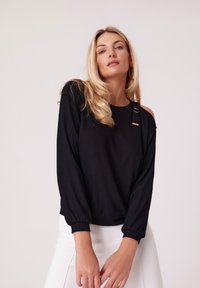 Czarny longsleeve z prążkowaną teksturą, z wycięciem na boku ramienia i metalowymi detalami. W połączeniu z białymi spodniami.