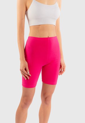 Kobieta ubrana w jasnoróżowe obcisłe krótkie spodenki motocyklowe i biały krótki top na tle jednolitego jasnego tła.