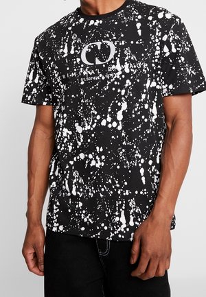 T-shirt noir avec motif d'éclaboussures de peinture blanche et texte blanc indiquant "CRIMINAL DAMAGE" ainsi que les coordonnées géographiques sur la poitrine.