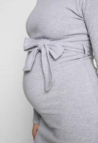 Vestido gris con ribetes que presenta un detalle de lazo lateral en la cintura, mangas largas y una silueta ajustada que resalta el vientre redondeado.