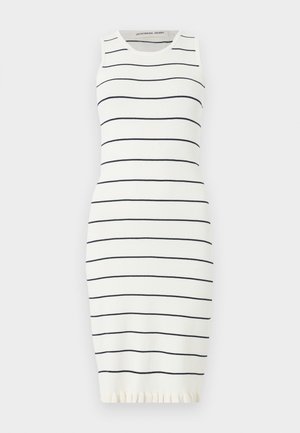 Γυναίκα στέκεται με το ένα χέρι στη μέση, φορά ένα λευκό αμάνικο φόρεμα με λεπτές μαύρες οριζόντιες ρίγες και μαύρα μυτερά παπούτσια χωρίς τακούνι.