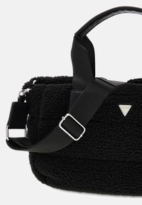 Czarna torebka o teksturowanej powierzchni, z miękką, puszystą fakturą, z czarnym paskiem i eleganckimi elementami metalowymi, obok trójkątnego logo.