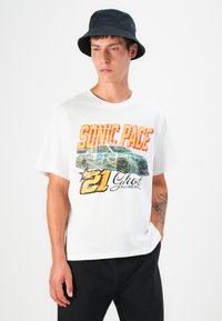 T-shirt graphique blanche avec un design de voiture de course coloré, affichant les textes "SONIC PACE" et "21 Ghost Worldwide" ; accompagné d'un chapeau seau noir.