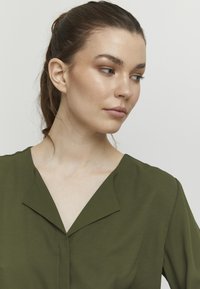 Green collared blouse made of lightweight fabric. Features a deep V-neckline and smooth texture. Minimalist design with no patterns or accents.