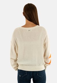 Maglione color crema con maniche lunghe, caratterizzato da accenti arancioni sulle maniche, orlo a costine e dettagli di cucitura visibili sulle spalle.