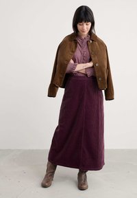 Brūns korduroja īsais žakets virs izsistā plūmes krāsas blūzes un ilga plūmes krāsas korduroja svārkiem. Brūnas potīšu zābaki papildina tērpu.