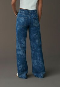 Vaqueros de mezclilla azul de pierna ancha con un patrón floral, que cuentan con bolsillos traseros y una cintura alta. La tela tiene una superficie suave y texturizada.
